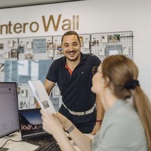 Eine Consultant sitzt am Schreibtisch vor ihrem PC mit Blättern in der Hand und unterhält sich mit einem Kollegen.